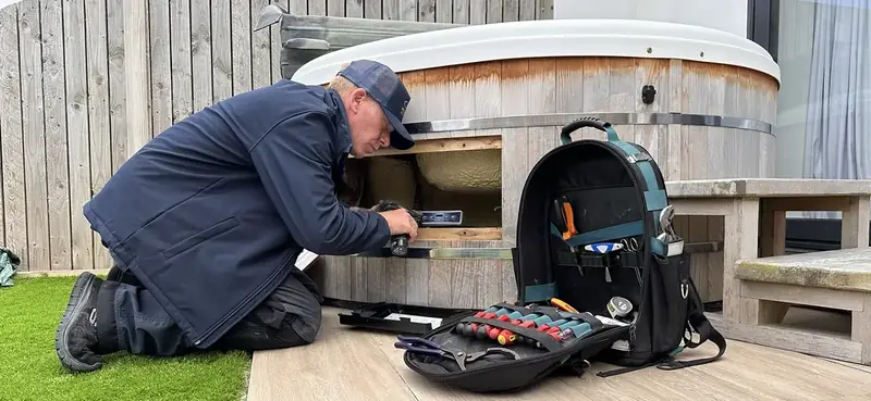 Jon kneeling at the open equipment bay of a round cedar hot tub, working on it with his tool bag beside him