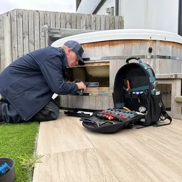 Jon kneeling at a hot tub with the cabinet panel open and his tool bag beside him