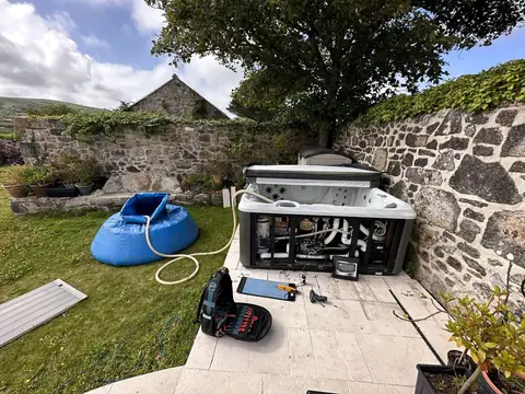Our blue holding bag filled with the tub’s water beside a hot tub in a walled garden, its side panel off