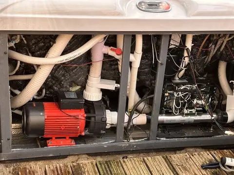 A hot tub with its cabinet panel off, showing a red pump, pipework and the control pack