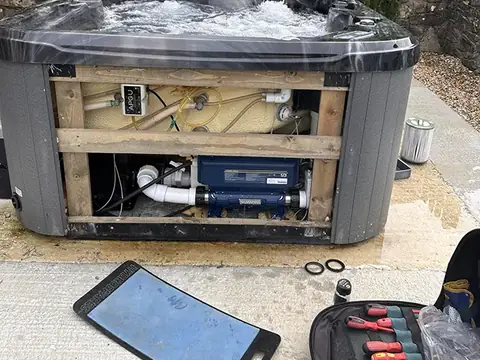 A grey hot tub with its side panel off and the equipment bay open, with a toolkit on the ground beside it