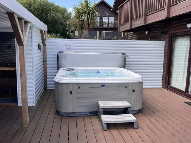 A holiday lodge hot tub with steps on a timber deck, its cover lifted and clear water, with lodges and a palm tree behind