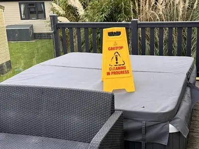 Covered hot tub on a lodge deck with a yellow “Caution: cleaning in progress” sign standing on the lid