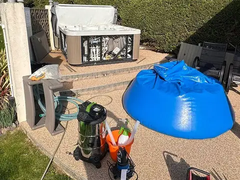 A hot tub with its cabinet open on a paved patio, our blue holding bag full of its water and a wet vacuum beside it