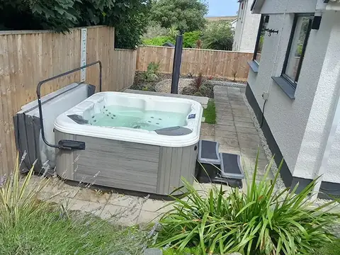 A hot tub with its cover off, set on paving against a wooden fence beside a house