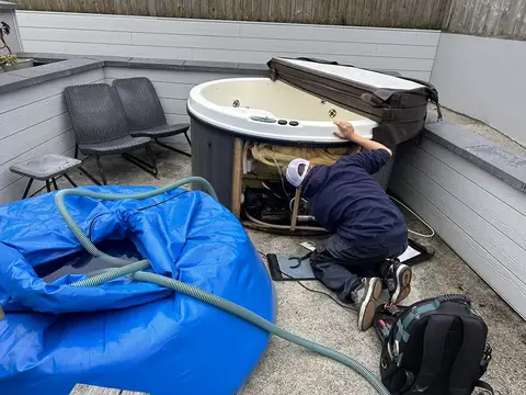 An engineer kneeling at an open hot tub cabinet in a walled courtyard, with the tub’s water held in our blue holding bag