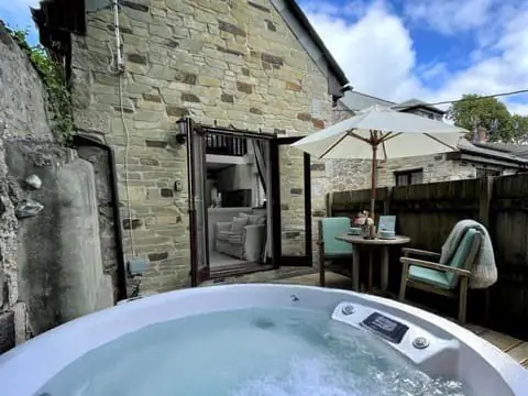 A hot tub in the courtyard of a stone cottage, with a parasol and garden chairs behind it