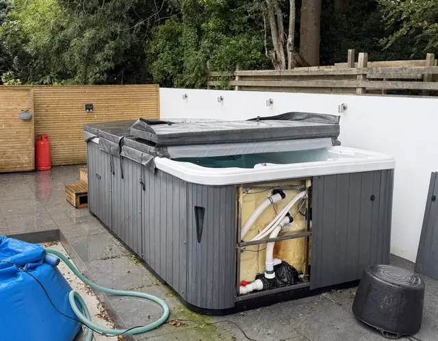 Long grey swim spa on a patio with one cabinet panel removed to show the pipework, its cover partly folded back, and a blue water bag and hose beside it
