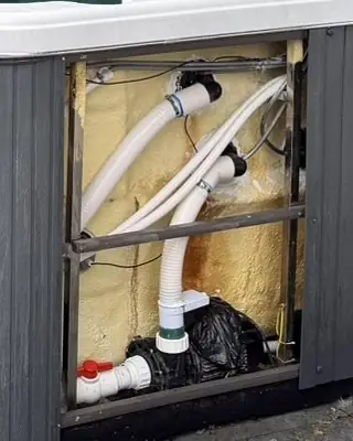 Inside a swim spa cabinet with the side panel off: white pipework and a valve, with insulation foam around them